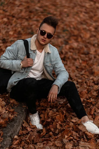 Cool joven estadounidense hipster en pantalones militares con estilo en una chaqueta de mezclilla de moda en gafas de sol negras en zapatillas blancas se sienta en el follaje seco dorado en un parque. Chico de moda se relaja al aire libre . — Foto de Stock