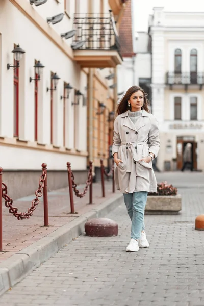 Modern European young woman in fashionable spring clothes in white stylish sneakers walks on the street near vintage buildings. Urban girl model travels on a road in the city. Casual youth fashion. — Stockfoto