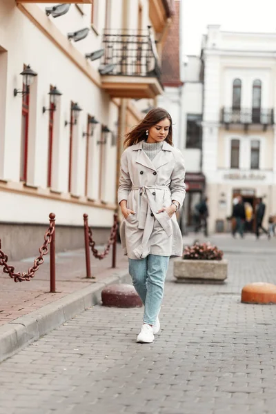 Stylish model of a beautiful young woman in blaue jeans in white shoes in a fashionable trench coat walks the street near vintage buildings. Attractive girl in trendy outerwear outdoors. Spring style. — Stockfoto