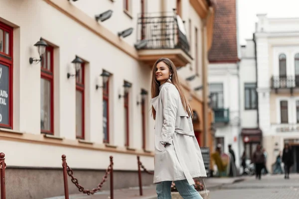 Happy elegant young woman model in a fashionable trench coat with chic brown long hair with natural make-up outdoors in the city. Joyful girl smiling and walks down the street. Spring style. — Stockfoto