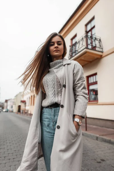 Modelo de mulher urbana jovem em um casaco de trincheira vintage em jeans azuis em um suéter elegante de malha está descansando na rua perto do edifício. Europeu bonito menina no outono-primavera roupas ao ar livre . — Fotografia de Stock