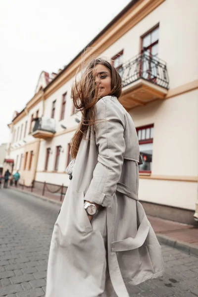 Utsikt från baksidan på en positiv attraktiv ung kvinna i en mode trenchcoat med lång chic brunt hår på en gata i staden. Vacker flicka reser genom stadens gator en varm höstdag — Stockfoto