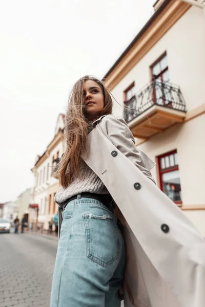 Söt stilren modell av en ung kvinna i trenchcoat i jeans i stickad vintagetröja går nerför gatan. Attraktiv europeisk stad flicka går och vänder tillbaka. Höst-vår mode. — Stockfoto