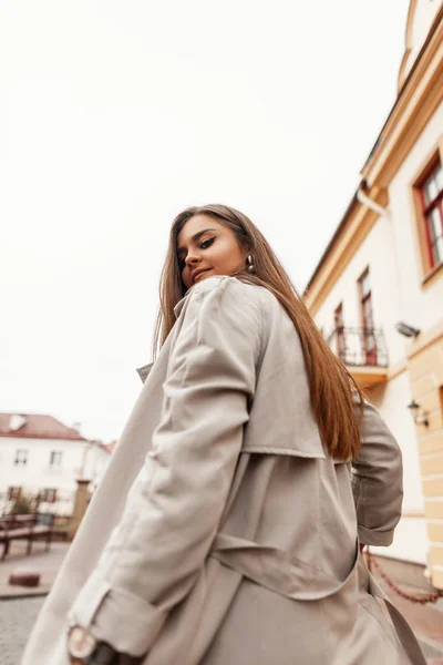 Ganska modern ung kvinna i en snygg trenchcoat går ner på gatan och vänder tillbaka. Modern trendig tjej modell poserar i staden på gatan. Våren damernas ytterkläder. — Stockfoto