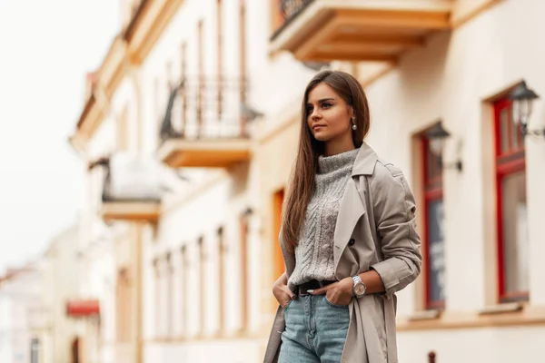 Modelo de uma mulher muito elegante em um casaco de trincheira bege chique com cabelo longo marrom em um suéter de malha elegante em um jeans anda pela cidade em um dia de primavera brilhante. Menina bonito moderno. Moda jovem . — Fotografia de Stock