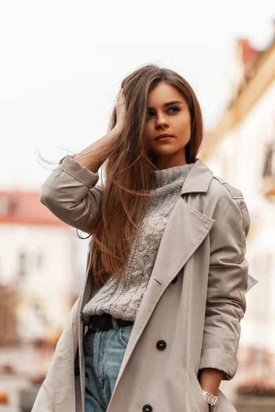 Hübsche urbane junge Frau glättet die Haare. schöne stilvolle Mädchen ein Mode-Modell in einem modischen Trenchcoat in einem Vintage-grauen Strickpullover geht durch die Stadt in der Nähe der Gebäude. Federstil. — Stockfoto
