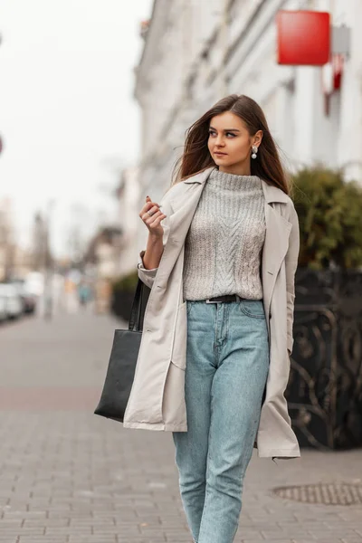 Modelo de moda de una mujer joven en una gabardina vintage en un suéter de punto elegante con una bolsa de cuero negro camina por la ciudad cerca de un edificio blanco. Linda chica de moda de cabello castaño viaja por la calle . — Foto de Stock