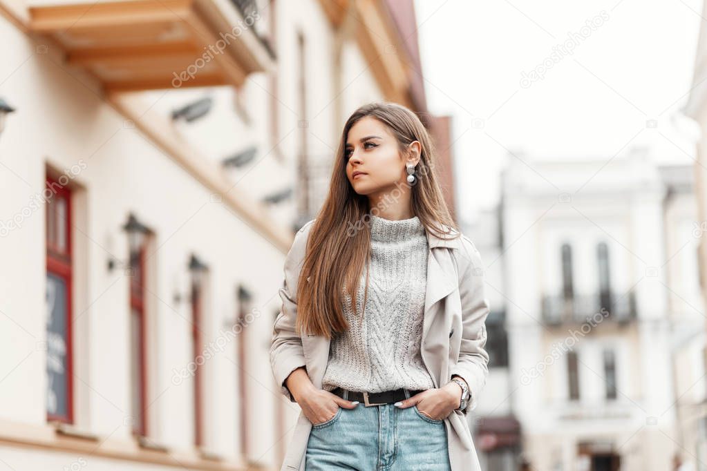Pretty attractive young woman in jeans in a trendy knitted sweater in a stylish beige trench coat walks on a street in the city near vintage buildings. Fashionable girl fashion model resting outdoors.