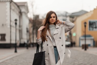 Moda örgü kazaklı, şık mavi kot pantolonlu, deri çantalı, şehirde taş bir yolda yürüyen zarif, modern bir genç kadın. Şirin kız mankeni sokaklarda geziyor..