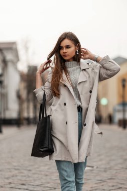Modaya uygun, uzun trençkotlu, mavi kot pantolonlu, şık, siyah deri çantalı güzel bir kadın şehirde cadde boyunca yürümenin keyfini çıkarıyor. Çekici moda kahverengi saçlı kız dışarıda..