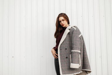 Kaliteli, zarif örgü süveterli kürklü, eski moda bir ceketle sokakta beyaz çizgili bir duvarın yanında duran güzel bir genç kadın modeli. Çekici modern kız şehirde poz veriyor..