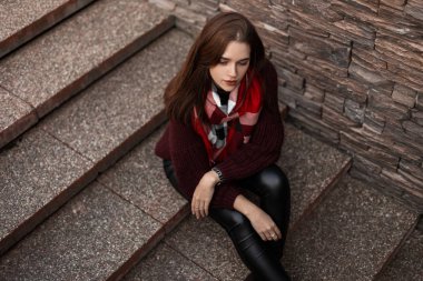 Havalı kıyafetler içinde güzel bir genç bayanın manzarası. Moda burgundy 'li çekici bir kız moda deri taytlı süveter örüp sokakta taş basamaklarda oturan bir eşarpla oturuyor.