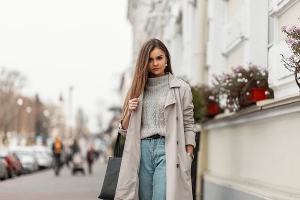 Mujer joven bastante elegante en un suéter de punto de moda en jeans en una gabardina de moda con una bolsa de pie en la calle cerca de un edificio blanco. Una chica atractiva camina por la calle. Estilo primavera . —  Fotos de Stock