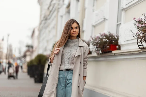 Jovem mulher bonita moderna em um suéter vintage em um casaco de trincheira na moda em jeans com um saco de couro preto em pé em uma rua na cidade em um dia quente de outono. Modelo menina muito chique caminha ao ar livre — Fotografia de Stock