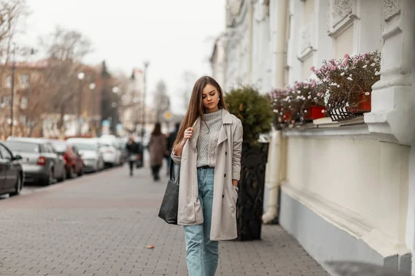 Jeune femme tendance dans un élégant trench-coat en pull chaud tricoté en jeans avec un sac en cuir noir à la mode près d'un bâtiment blanc dans la ville. Fille urbaine à la mode à l'extérieur. Mode des jeunes femmes . — Photo