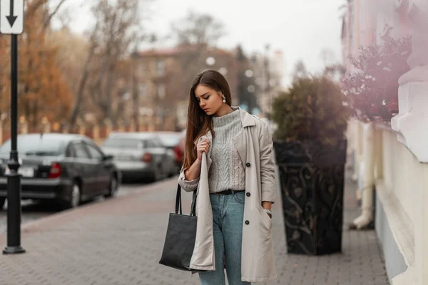 Mulher nova bonita em um suéter em um casaco de trincheira leve longo na moda em calça com uma bolsa de couro preta que está em uma rua na cidade em um dia de outono quente. Bonita modelo menina urbana ao ar livre . — Fotografia de Stock