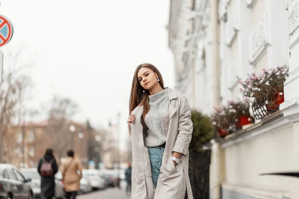 Vacker modern modell av en ung kvinna går i en snygg trenchcoat i en stickad tröja i jeans på staden. Attraktiv tjej i trendiga kläder nära en vintage byggnad på gatan. Tillfällig stil — Stockfoto