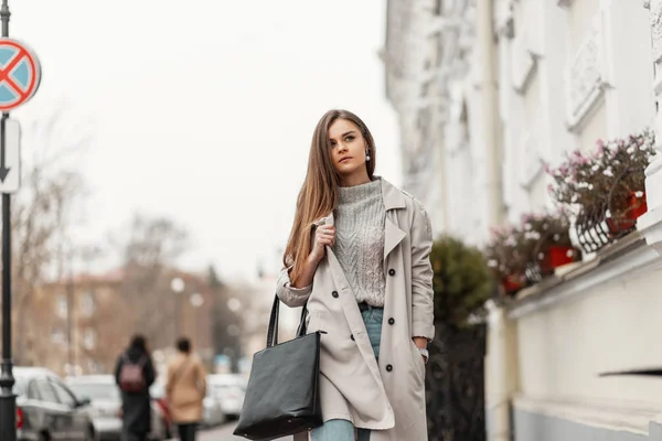 Modelo de uma jovem mulher de cabelos castanhos em um casaco de moda em uma camisola de malha elegante com uma bolsa preta de couro caminha na cidade perto de um edifício branco. Menina da moda atraente viaja pela rua . — Fotografia de Stock