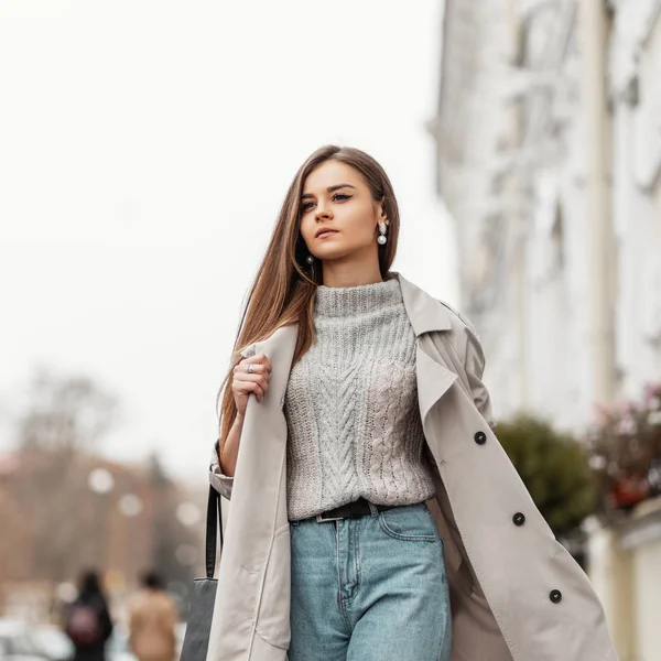 Uma jovem muito elegante anda ao ar livre na cidade. Linda modelo menina de cabelos castanhos em outerwear elegante com um saco de couro viaja ao longo da rua. Roupas femininas da Primavera. Estilo casual . — Fotografia de Stock