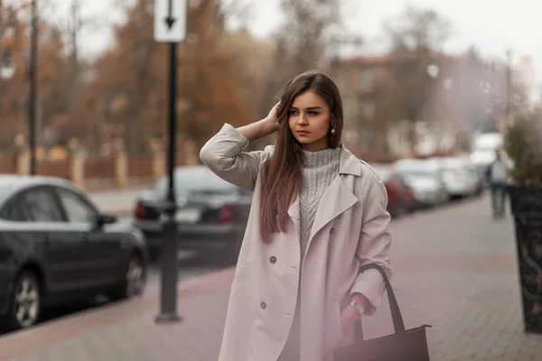 Aranyos fiatal nő hosszú barna haj egy elegáns könnyű ballon egy divatos fekete bőr táska áll az utcán. Vonzó lány modell sétál a városban egy meleg őszi napon. — Stock Fotó