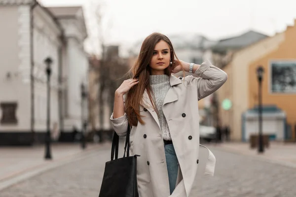 Jolie jeune femme moderne dans un pull tricoté à la mode en jeans bleu tendance dans un élégant trench coat avec un sac noir en cuir marche sur une route en pierre dans la ville. Modèle fille mignonne voyage dans la rue . — Photo