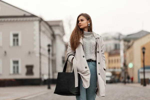 Jovem encantadora em uma camisola de malha elegante em um casaco de trincheira chique em jeans azuis com uma bolsa preta está de pé em uma rua na cidade. Menina de cabelos castanhos atraente ao ar livre. Moda casual . — Fotografia de Stock