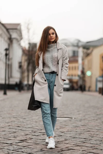 Jovem atraente de cabelos castanhos em roupas de primavera na moda em tênis elegantes brancos com um saco de couro caminha ao longo da rua perto de edifícios vintage. Menina bonito caminha em uma estrada de pedra na cidade — Fotografia de Stock