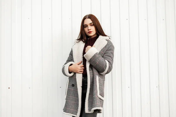 Beautiful model of an attractive young woman in a vintage checkered jacket with fur in a stylish knitted sweater stands in the city near a white striped wall.Cute brown-haired girl poses on the street — Stock Photo, Image