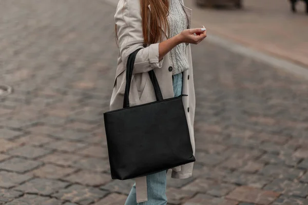 Corpo de close-up de uma jovem mulher em uma camisola de malha vintage em um elegante casaco de trincheira com um elegante saco de couro preto na cidade. Coleção outono-primavera de outerwear feminino na moda e acessórios . — Fotografia de Stock