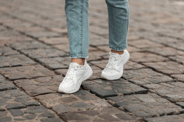 Modern young woman in a stylish blue jeans in fashionable white sneakers stands on a stone road in the city. Stylish women's shoes. Youth style. Closeup of female legs in a trendy shoes.