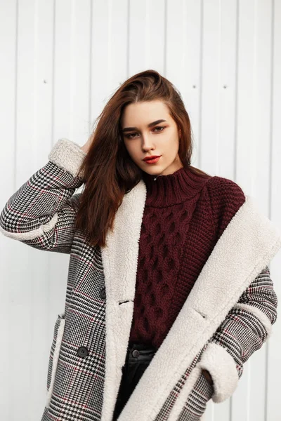 Model of a attractive cute young woman in a stylish knitted burgundy sweater in a checkered warm long jacket with fur posing near a white wall on the street. Beautiful fashionable girl outdoors. — Stock Photo, Image