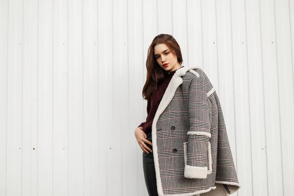 Lindo modelo de una hermosa mujer joven en una chaqueta a cuadros vintage con piel en un suéter de punto con estilo cálido se encuentra en la calle cerca de una pared de rayas blancas. Atractiva chica moderna posa en la ciudad . — Foto de Stock