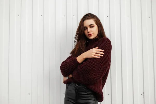 Aantrekkelijk model jonge vrouw in stijlvol lederen zwarte broek in een modieuze gebreide trui poserend in de buurt van een moderne witte muur op straat. Mooi bruinharig meisje buiten. — Stockfoto