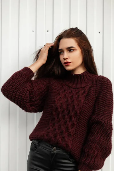 Attractive portrait of a young stylish woman in a vintage burgundy knitted sweater with brown hair with natural makeup of a near white wall. Beautiful fashionable girl model posing near a building. — Stock Photo, Image