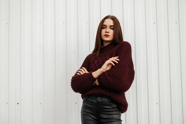 Modèle d'une belle jeune femme dans un élégant pull tricoté bordeaux en cuir pantalon noir tendance posant dans la rue près d'un bâtiment blanc. Fille urbaine à la mode dans la ville un jour d'automne . — Photo