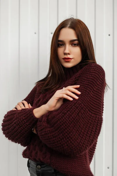 Beautiful portrait of a young stylish woman in a vintage burgundy knitted sweater with brown hair with natural makeup of a near white wall. Attractive fashionable girl model posing near a building. — Stock Photo, Image