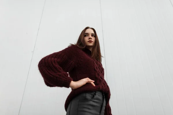 Modelo de uma jovem na moda em uma camisola elegante borgonha em couro calças pretas na moda posando na rua perto de um edifício branco. Menina urbana bonita na moda na cidade em um dia de outono . — Fotografia de Stock