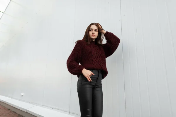 Evropská hezká mladá módní žena ve stylovém burgundském svetru v módních černých kožených kalhotách pózujících ve městě u bílé zdi. Atraktivní krásná dívka model venku. Mládež oblečení. — Stock fotografie