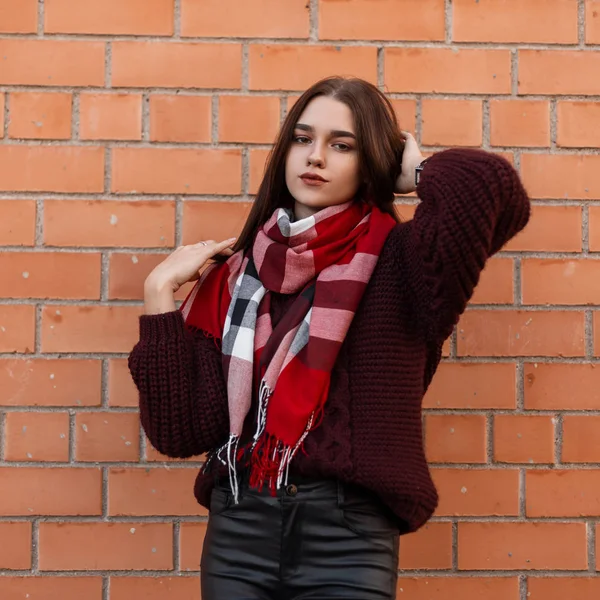 Modèle d'une jolie jeune femme aux cheveux bruns dans un pull tricoté à la mode avec une écharpe à carreaux en pantalon de cuir dans la ville près d'un mur de briques. Élégante fille mignonne se repose à l'extérieur dans la rue . — Photo