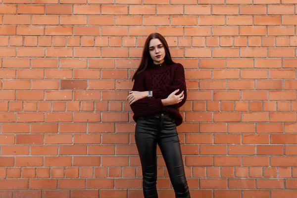 Evropská hezká mladá módní žena ve stylovém pleteném burgundském svetru v módních černých kožených kalhotách stojí ve městě u cihlové zdi. Městská krásná dívka modelka venku. Mládež oblečení. — Stock fotografie