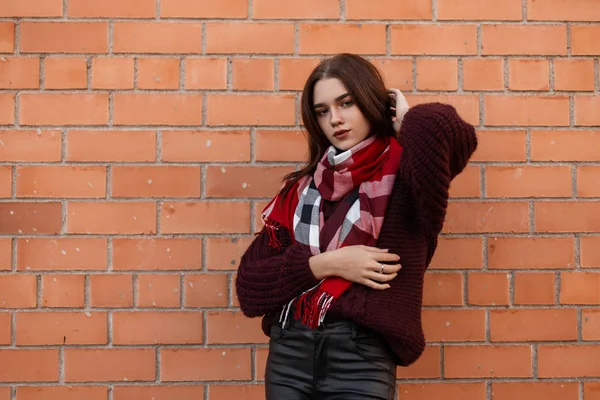 Jeune femme élégante dans un pull tricoté bordeaux à la mode avec une écharpe rouge à la mode dans un pantalon en cuir posant à l'extérieur près d'un bâtiment en brique. Jolie belle fille se détend dans la ville. Mode d'automne . — Photo