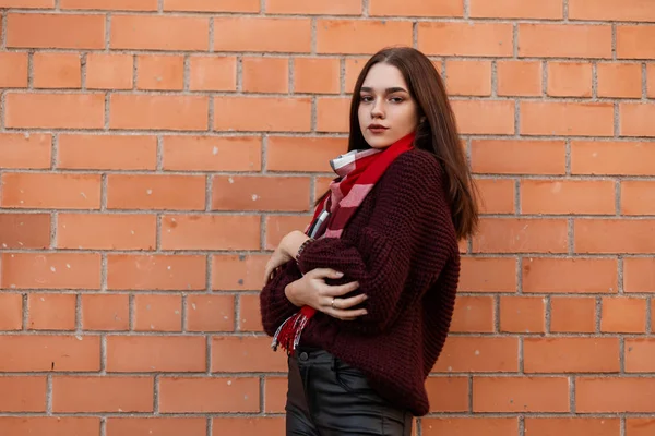 Mignonne jolie jeune femme dans un élégant pull tricoté avec une écharpe à carreaux rouges de mode en pantalon en cuir pose près d'un bâtiment en brique vintage dans la rue. Européen modèle de fille à la mode en plein air — Photo