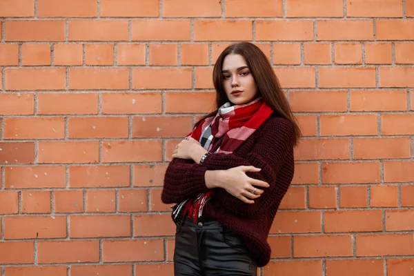 Jolie jeune femme urbaine dans un pull bordeaux mode en pantalon en cuir noir à la mode avec une écharpe à carreaux posant dans la ville près d'un mur de briques. Modèle fille moderne à l'extérieur. vêtements de mode jeunesse . — Photo