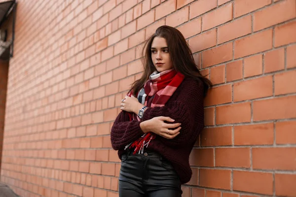 Jolie jolie jeune femme dans un élégant pull tricoté avec une écharpe à carreaux rouges à la mode dans un pantalon en cuir se repose près d'un bâtiment en brique vintage dans la rue. Belle fille à la mode en plein air . — Photo