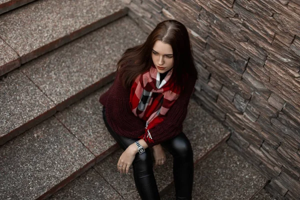 Mulher bonita europeia em uma camisola borgonha de malha vintage em calças de couro preto na moda com um lenço xadrez senta-se em uma escada de pedra. Modelo de menina urbana na moda está descansando na rua . — Fotografia de Stock
