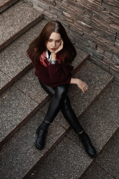 Moderno joven en botas en un suéter vintage de punto de moda en pantalones de cuero con una bufanda a cuadros está sentado en la ciudad en una escalera de piedra. Hermosa chica en ropa de moda está descansando al aire libre . — Foto de Stock