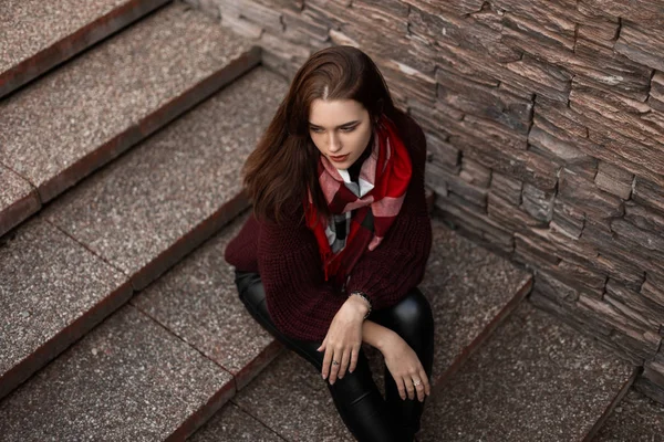 Jeune belle femme dans un pull vintage tricoté à la mode en pantalon en cuir avec une écharpe à carreaux est assis dans la ville sur un escalier en pierre. Fille attrayante dans des vêtements à la mode se repose à l'extérieur . — Photo