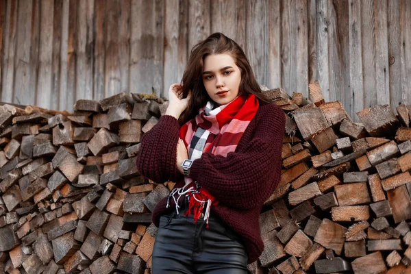 Modelo bonito de uma jovem mulher rural em uma camisola de malha elegante com um lenço na moda fica perto de uma antiga parede de madeira com troncos. Menina bonita descansando na aldeia em um dia quente de outono . — Fotografia de Stock