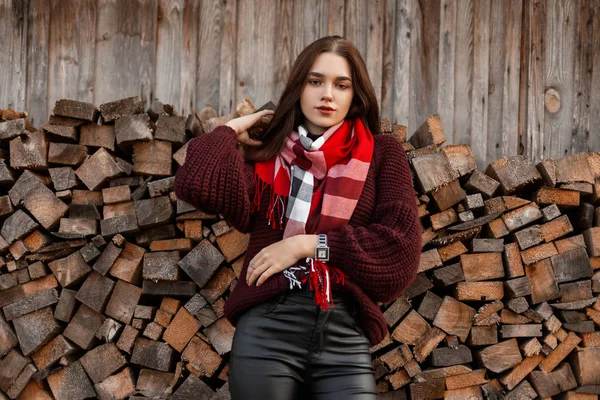 Modell av en söt ung kvinna i en vintage stickad tröja med en varm rutig trendig halsduk i läderbyxor poserar nära en gammal by skjul. Attraktiv ganska snygg flicka står nära stockar utomhus — Stockfoto