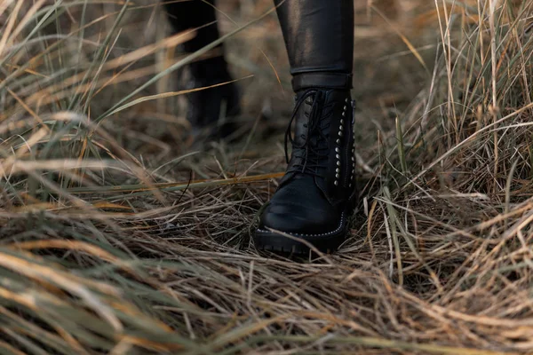 Moderne jonge vrouw in zwarte stijlvolle legging in modieuze leren laarzen staat op het droge gras in het park. Trendy collectie damesherfstschoenen. Close-up van vrouwelijke benen. — Stockfoto